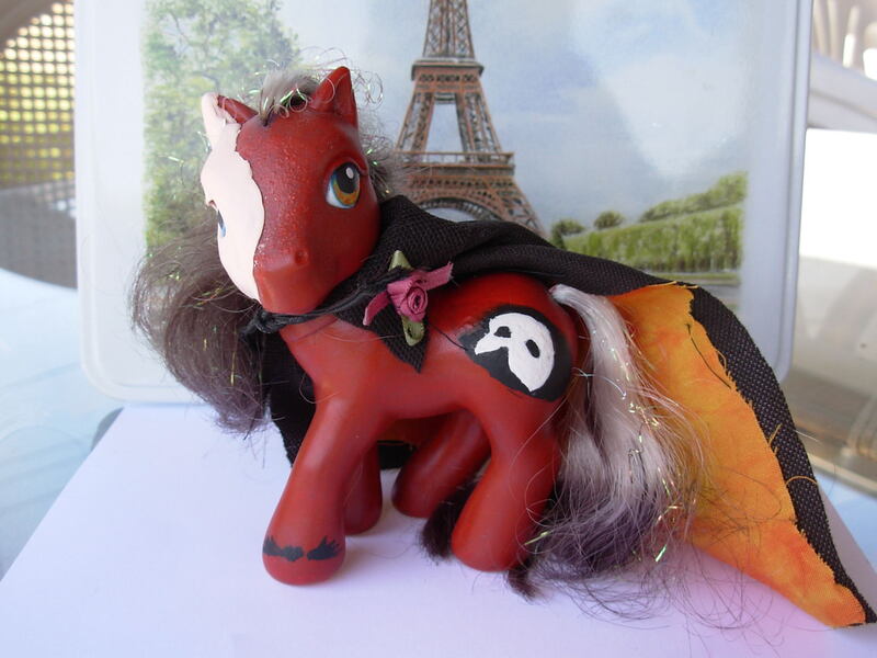 A red G3 MLP with a sculpted white half mask, one orange and one blue eye, black and white mane and tail, and a Phantom of the Opera mask logo as his cutie mark; he is wearing his accessory, a black cape with an orange underside